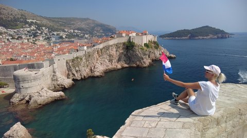 Woman with Croatian flag on Fort Lovrijenac fortress looking walls of Dubrovnik of Croatia. Over the West Harbour of Dubrovnik historic town with Fort Bokar of Croatia in Dalmatia. UNESCO walled city