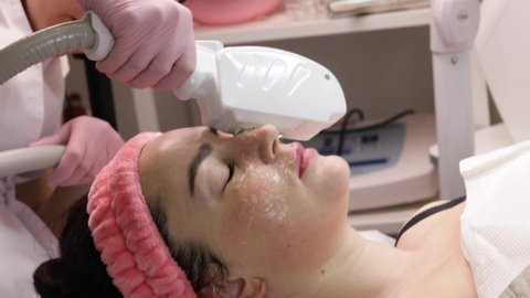 Smiling woman enjoying ELOS skin rejuvenation facial. Female with cooling gel on face and laser protection glasses, lying therapy table at beauty clinic, treating skin from blemishes, doing facelift.