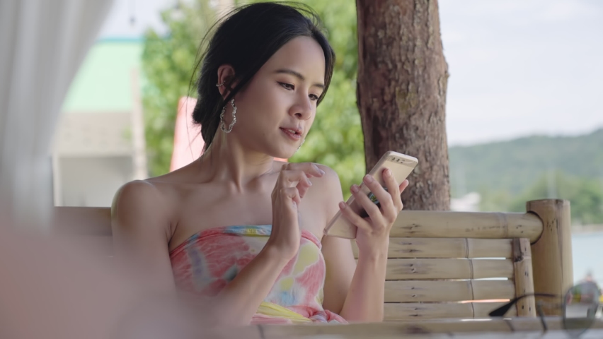Asian sexy young tourist using sitting on beach bamboo chair using smartphone, technology woman using finger on touch screen phone, distance communication helper, online booking reservation 