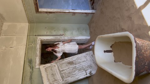 Vertical video. Young woman in robe in abandoned building in bathroom walks bath tub. Ghost town Kolmanskop in desert. Room filled with sand. Old bathtub in ruined house. Girl in white silk underwear