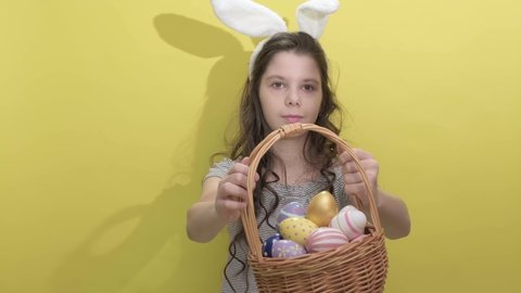 4k. Happy easter. Beautiful cute little girl in Easter bunny ears holds a basket with eggs on a yellow background. Easter activities for children. Hello spring and easter concept