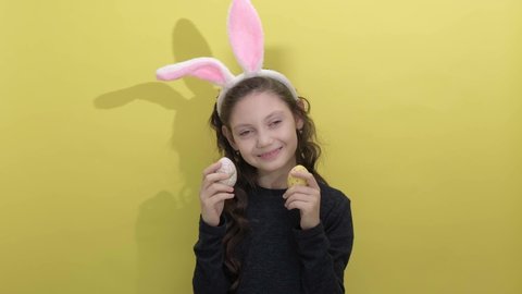 4k. Happy easter. Beautiful cute little girl in Easter bunny ears holding two painted easter eggs on a yellow background. Easter activities for children. Hello spring concept