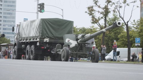 Minsk, Belarus - June 28 2018: Column of military trucks transports large-caliber cannons along the city street. People stand and look along road on cloudy day. 
