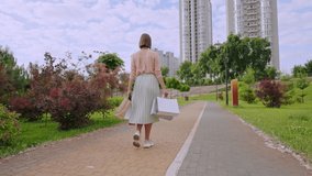 rear caucasian woman holding varicoloured paper bags walking in city park slow motion