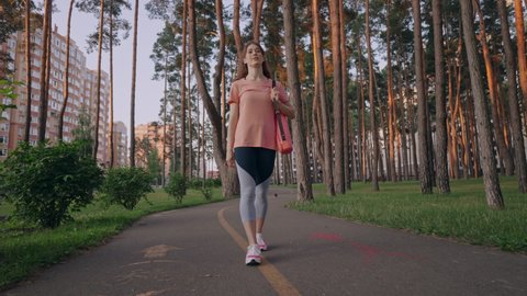 happy young woman with yoga mat wearing sportswear walking in city park with pines active sport healthy lifestyle