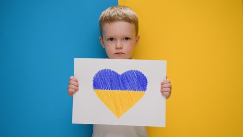 Blond boy holding banner with yellow-blue heart drawn with marker in banner. Kid will support Ukraine by showing heart on yellow-blue flag. Ukrainian crisis, Russian aggression in Ukraine.