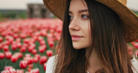 Portrait Face of Romantic Caucasian Girl standing on the Field with Tulips. Woman in Straw Hat enjoys Nature and Flowers Outdoors. Having Natural Face. Blue, Azure Eyes. Long Hair. Spring.