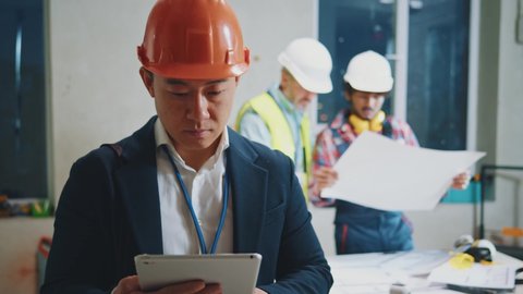 Serious portrait asian engineer, architect wearing a white protective helmet in a suit use tablet computer stand. On bakground builders in hardhats working in building. Slow motion