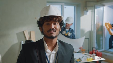 Handsome young engineer, foreman, architect wearing a white protective helmet in a suit look at camera. On bakground builders in hardhats working in building. Slow motion