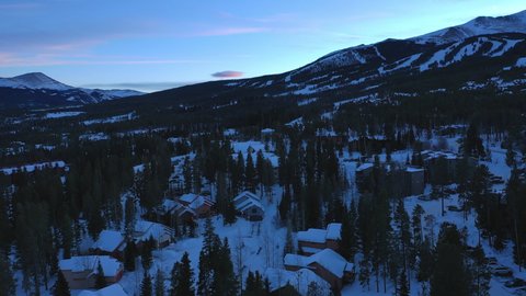 Aerial drone shot over buildings and houses in Breckenridge, Colorado, United States at sunset over mountainous terrain covered with trees during winter season.