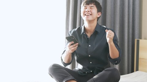Young asian man feeling happy and excited  ed expression after seeing the good news from smartphone, sit down on the bed on sunny day, winning big jackpot prize, success taking benefit from investment