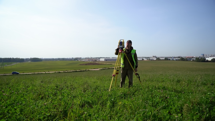 A surveyor in a reflective vest, taking measurements with a total station, waving to his assistant. Surveying in the field, warm season, topography and geodesy. General plan. Royalty-Free Stock Footage #1088076433