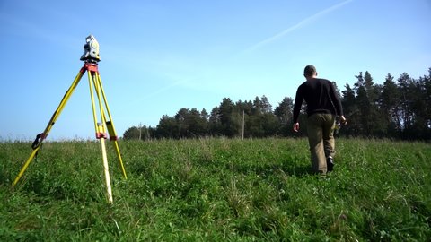 A surveyor with a reflector in his hand is moving away from the total station standing on a tripod in the field and walking towards the point to be measured. A professional surveying engineer surveys.