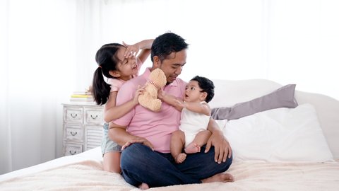4K 50fps, Asian father holding an adorable newborn boy playing brown teddy bear on the bed, and an older sister hugging dad behind him playing with his younger brother, happy face.Family and Healthy.