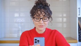 Beautiful white woman doing video call with mobile phone. Young adult female in glasses speaking on frontal camera of modern blue smartphone. 4k video of brunette with curly hair talking on video