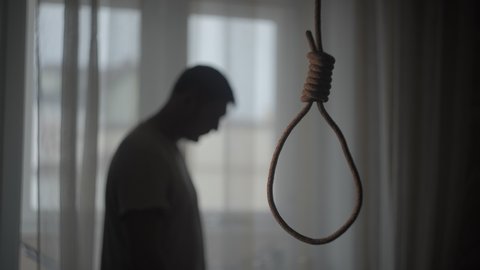 A rope noose hangs in a dark apartment. The man is depressed and upset. He is contemplating suicide. The concept of personality crisis.