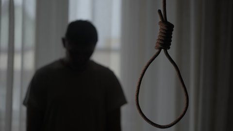 A rope noose hangs in a dark apartment. The man is depressed and upset. He is contemplating suicide. The concept of personality crisis.