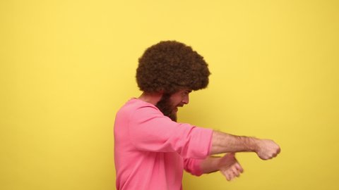 Side view of bearded man with Afro hairstyle pretending to hold rope in hands and pull, expressing much effort on face, wearing pink sweatshirt. Indoor studio shot isolated on yellow background.