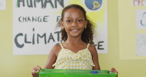 Video of happy african american girl holding box with recycling symbol in classroom. primary school education and learning concept.