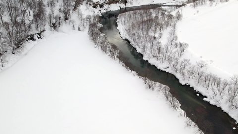 Beautiful clear river with meltwater flowing gently between the thin trees covered in a thick layer of snow Hokkaido Province in Japan. High angle drone dolley shot