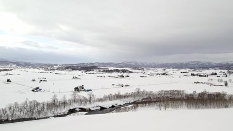 Flat farmlands with large farms covered in a thick layer of white snow with the high mountains of Hokkaido in Japan in the background. High angle drone dolley shot