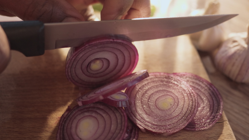 Macro shot of African American male hands chopping a raw red purple onion with sharp kitchen knife on a thin rings.