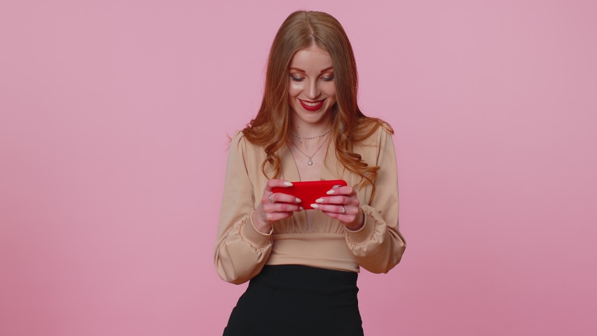 Worried funny addicted woman enthusiastically playing racing video games on mobile phone. Young adult teen girl using smartphone gadget app with drive simulator isolayed on pink studio wall background