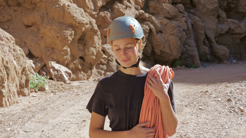 Rock climber in a helmet walks along a canyon, holds rope. Sportive girl smiling, keeping climbing equipment. Female mountaineer is preparing to climb mountain. Alpinist woman near cliff
