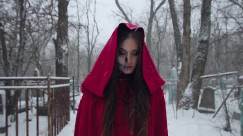 Young woman with scary face in red hood stands in the daytime in cemetery in winter. Little Red Riding Hood with skeleton makeup in halloween 