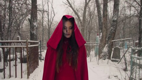 Evil spirits in red cloak and hood in cemetery in winter. Beautiful young woman with halloween makeup. Grim with skeleton teeth
