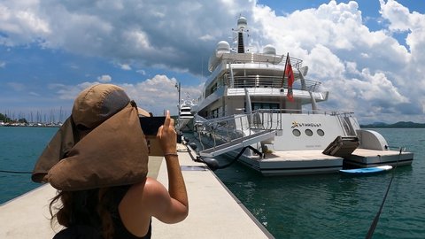 Phuket, Thailand, 02, March, 2022:
A girl photographs a superyacht in a yacht club, a girl in a headdress photographs a superyacht, walking along the yacht marina