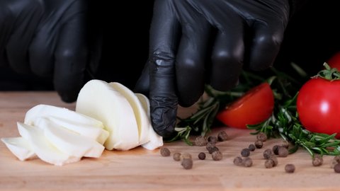 Female chef cutting fresh mozzarella cheese in slices for caprese salad with tomatoes. Hands slice cheese with kitchen knife. Mozzarella on cutting board, cookware and ingredients on kitchen table