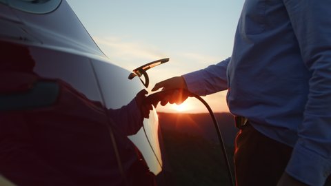 Male hand inserts power connector into EV car. Unrecognizable man plugging in charging cable to to electric vehicle and charges batteries with golden sunset on cinematic background, RED camera shot 6K