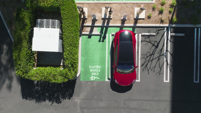 Aerial of electric car vehicles recharging. EV eco friendly electricity powers cars. Drone descending above woman plugging in charging cord in red SUV sedan electric vehicle at summer, RED camera Royalty-Free Stock Footage #1088106445
