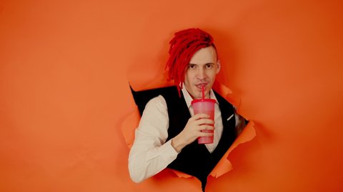 Barman with drink looking through hole in paper background. Male bartender with red dreadlocks and cup of sweet beverage peeking through ripped orange paper background in studio and looking at camera