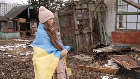 a girl wearing in the Ukrainian flag looks at the destroyed houses. War in Ukraine