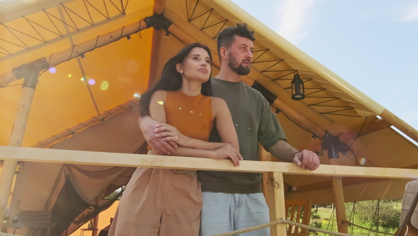 Low angle medium long of young loving hetero couple hugging, talking and standing by fencing on terrace of summer house on sunny day