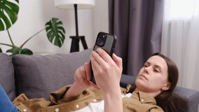 Selective focus of young woman lying on sofa in living room browsing surfing wireless internet on smartphone, happy girl rest on couch at home message text on modern cellphone. Shopping online