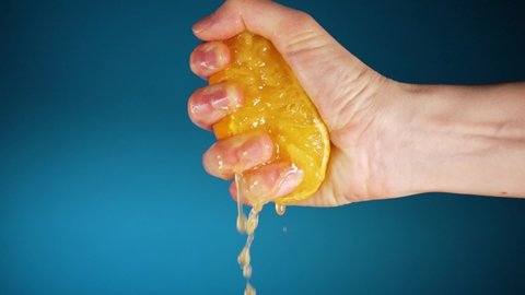 Woman's hand squeezes half a juicy orange on a blue background in slow motion. The juice slowly flows down the fingers.