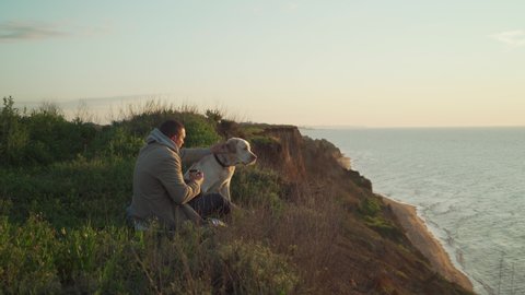 Young attractive male caress his retriever dog sitting on a cliff near sea