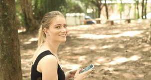 Young woman using a smartphone at day time with a green park. High quality video. Mobile phone, technology, urban concept. 