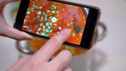 A Woman Looks at a Photo of Traditional Ukrainian Red Borscht on a Smartphone. Photo of food through the camera of a mobile phone in a mobile application. Home kitchen. Lunchtime.