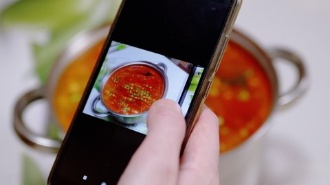 A Woman Looks at a Photo of Traditional Ukrainian Red Borscht in a Smartphone. Photographing food. Female Hands view photos of food on the phone screen mobile phone in an application. Home kitchen.