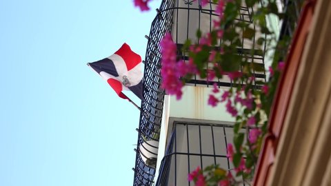 Slow motion of the Dominican Republic flag waving in the wind against the blue sky. Pink Bougainvillea flowers on the balcony of the building in the Colonial City