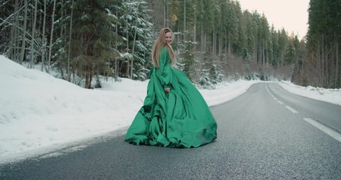 Caucasian blonde woman long hair walking running on winter snowing asphalt road in green dress brutal shoes. Fantasy girl runs winter forest. long vintage dress fabric waving. Slow motion 120 fps 4k