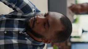 Vertical video: Portrait of african american person smiling in office, sitting on couch to work on laptop. Company employee using financial statistics to create job development and business growth.