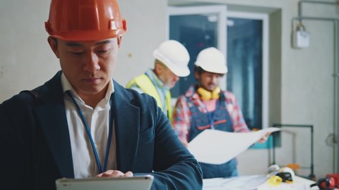 Asian engineer, architect wearing a white protective helmet in a suit use tablet computer stand look at camera. On bakground builders in hardhats working in building. Portrait. Slow motion