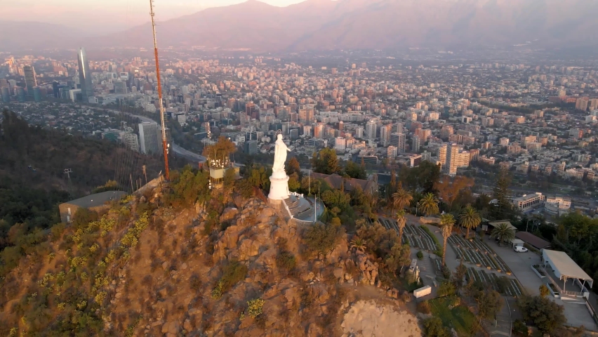 Aerial pan left of statue in Sanctuary of the Immaculate Conception in San Cristobal Hill summit, Santiago city in background at sunset, Chile