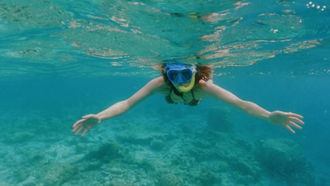 Woman with long loose dark hair in bikini and goggles shows peace sign swimming above coral reef in transparent ocean water slow motion underwater split shot