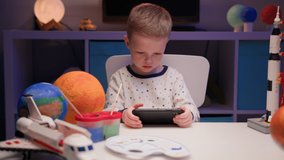 Little blond boy is playing video games on smartphone sitting at home at table in evening around many planets of solar system.